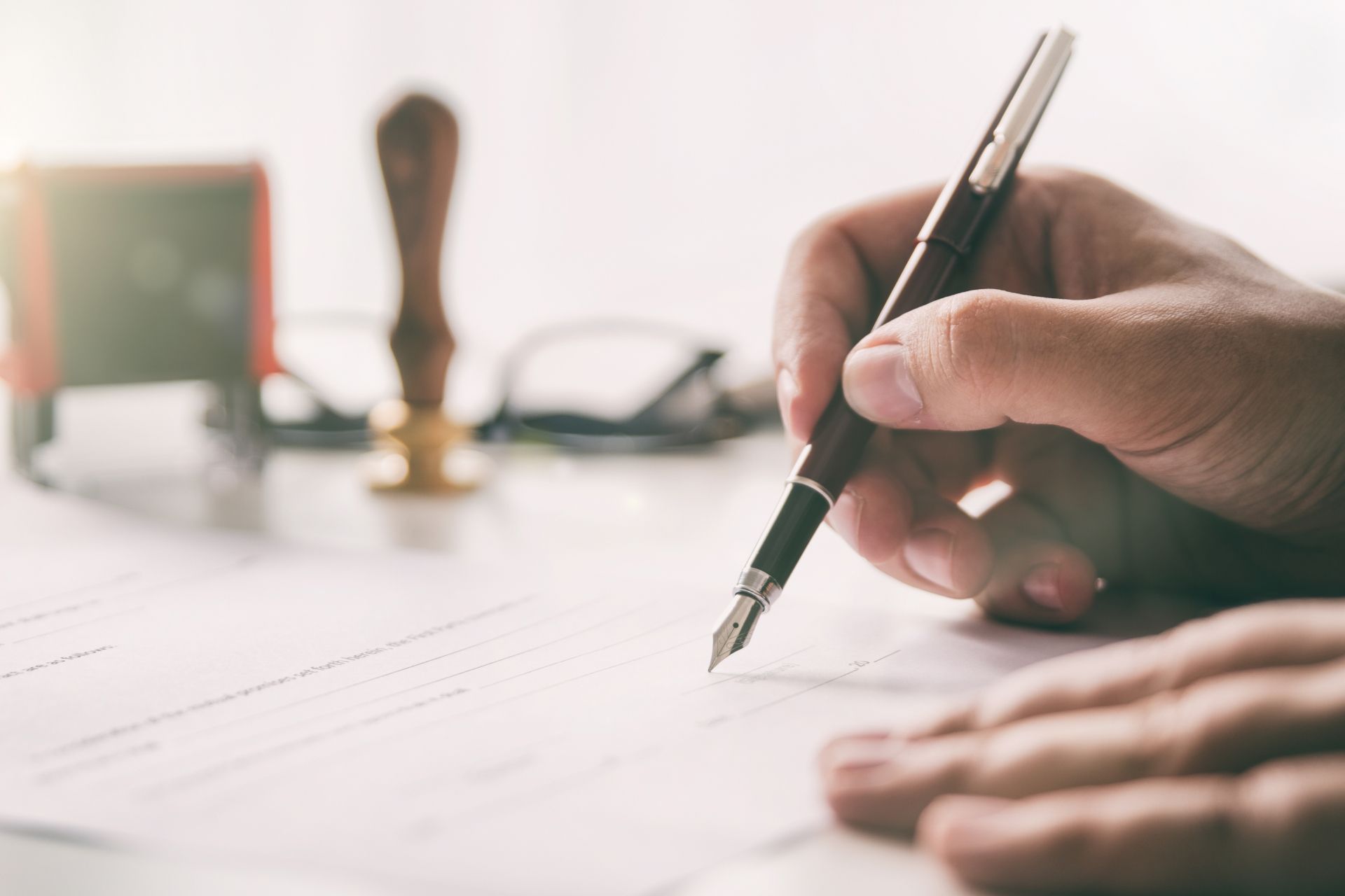 Männerhand unterschreibt ein Schriftstück mit Federfüllhalter, im Hintergrund befinden sich Stempel